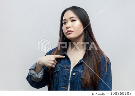 Portrait of brunette woman in blue denim jacket standing points at herself, proud of own achievements, looks confidently and self assured at camera. Indoor studio shot isolated on gray background. 108269108