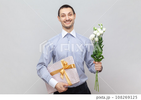 Smiling happy positive man standing with present box and bouquet of flowers, congratulate her girlfriend with birthday, wearing light blue shirt. Indoor studio shot isolated on gray background. 108269125