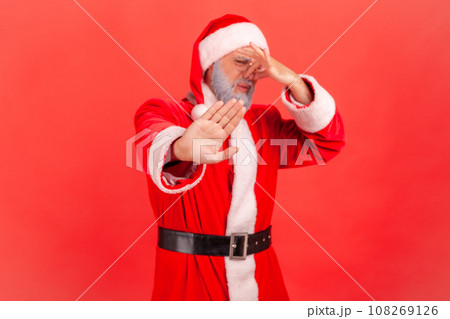 Santa claus in red costume pinching nose with fingers to avoid bad smell and showing stop gesture, demonstrating repulsion to stink, awful odor. Indoor studio shot isolated on red background. Santa claus in red costume pinching nose with fingers to avoid bad smell and showing stop gesture, demonstrating repulsion to stink, awful odor. Indoor studio shot isolated on red background. 108269126