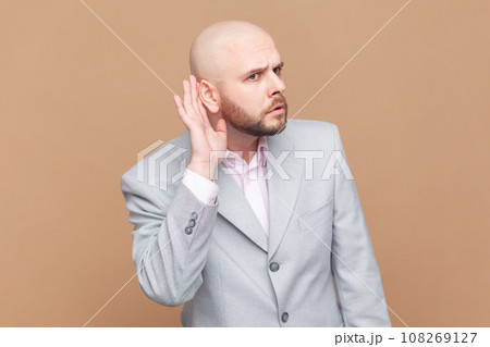 Portrait of bald man overhearing, listening intently to secret information, private talk, holding hand near ear to hear better, wearing gray jacket. Indoor studio shot isolated on brown background. Portrait of bald man overhearing, listening intently to secret information, private talk, holding hand near ear to hear better, wearing gray jacket. Indoor studio shot isolated on brown background. 108269127