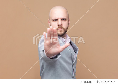 Portrait of bald bearded man does stop sign with palm, demonstrates prohibition and restriction, refuses something, says no, wearing gray jacket. Indoor studio shot isolated on brown background. Portrait of bald bearded man does stop sign with palm, demonstrates prohibition and restriction, refuses something, says no, wearing gray jacket. Indoor studio shot isolated on brown background. 108269157