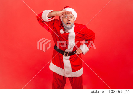 Elderly man with gray beard in santa claus costume keeping palm over head and looking attentively far away, peering with expectation at long distance. Indoor studio shot isolated on red background. Elderly man with gray beard in santa claus costume keeping palm over head and looking attentively far away, peering with expectation at long distance. Indoor studio shot isolated on red background. 108269174