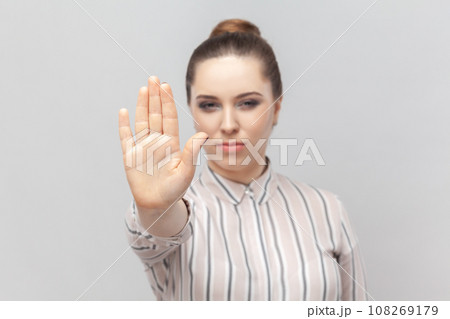 Portrait of young adult strict serious woman wearing striped shirt standing showing prohibition gesture with her palm, asking to stop. Indoor studio shot isolated on gray background. Portrait of young adult strict serious woman wearing striped shirt standing showing prohibition gesture with her palm, asking to stop. Indoor studio shot isolated on gray background. 108269179