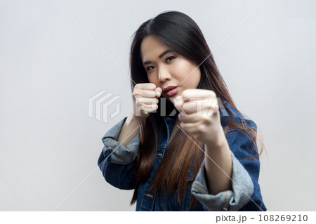 Portrait of angry aggressive beautiful serious brunette woman in blue denim jacket standing with clenched fists, fighting with somebody. Indoor studio shot isolated on gray background. Portrait of angry aggressive beautiful serious brunette woman in blue denim jacket standing with clenched fists, fighting with somebody. Indoor studio shot isolated on gray background. 108269210