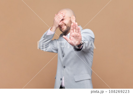 Portrait of scared frighten bald bearded man covering his face and showing stop hand gesture, sees something shameful, wearing gray jacket. Indoor studio shot isolated on brown background. 108269223