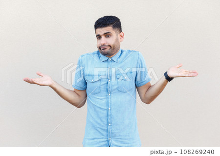 Portrait of confused uncertain man wearing denim shirt standing shrugging shoulders, doesn't know how to make a decision, looks doubtful. Indoor studio shot isolated on gray background. Portrait of confused uncertain man wearing denim shirt standing shrugging shoulders, doesn't know how to make a decision, looks doubtful. Indoor studio shot isolated on gray background. 108269240