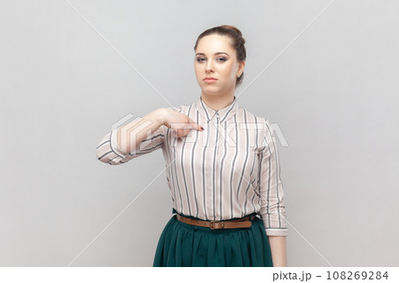 Portrait of strict bossy serious woman wearing striped shirt and green skirt standing pointing at herself with proud expression, looks arrogant. Indoor studio shot isolated on gray background. 108269284
