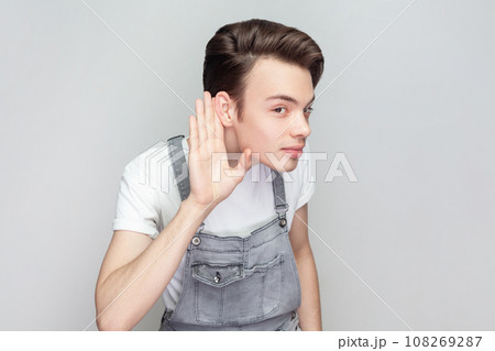 Intrigued curious young brunette man standing keeps hand near ear tries to overhear interesting conversation, looks attentively, wearing denim overalls. Indoor studio shot isolated on gray background. 108269287