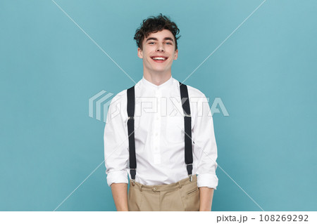 Portrait of joyful cheerful attractive young man wearing white shirt and suspender, expressing positive emotions, being in good mood. Indoor studio shot isolated on blue background. 108269292