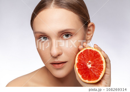 Closeup portrait of beautiful woman with perfect skin model looking at camera holding half of grapefruit. Indoor studio shot isolated over gray background. 108269314