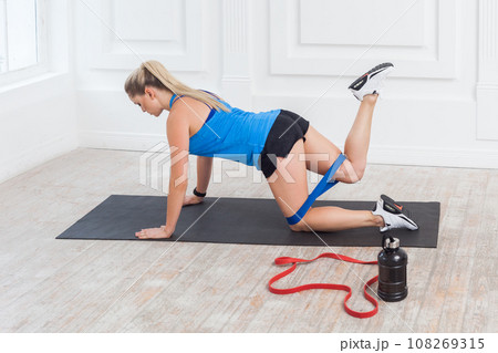 Side view portrait of athletic sporty beautiful young woman training with bands, trains legs and buttocks, fitness exercising with expander on yoga mat. Indoor studio shot. Side view portrait of athletic sporty beautiful young woman training with bands, trains legs and buttocks, fitness exercising with expander on yoga mat. Indoor studio shot. 108269315