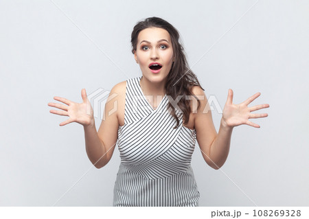 Portrait of woman exclaims happily, keeps palms raised, feels very happy, reacts on awesome news, gets pleasant surprise, wearing striped dress. Indoor studio shot isolated on gray background. Portrait of woman exclaims happily, keeps palms raised, feels very happy, reacts on awesome news, gets pleasant surprise, wearing striped dress. Indoor studio shot isolated on gray background. 108269328