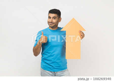 Portrait of delighted unshaven man wearing blue T- shirt standing holding carton arrow indicating up, being glad of career success, showing thumb up. Indoor studio shot isolated on gray background. 108269340