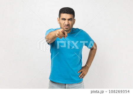 Portrait of serious unshaven man wearing blue T- shirt standing with hand on hip, pointing finger at camera, expressing negative emotions. Indoor studio shot isolated on gray background. Portrait of serious unshaven man wearing blue T- shirt standing with hand on hip, pointing finger at camera, expressing negative emotions. Indoor studio shot isolated on gray background. 108269354