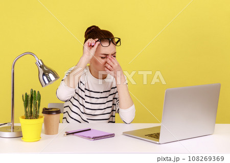 Exhausted stressed woman office worker rubbing tired of laptop work eyes, putting off glasses, sitting workplace, overworking. Indoor studio studio shot isolated on yellow background. Exhausted stressed woman office worker rubbing tired of laptop work eyes, putting off glasses, sitting workplace, overworking. Indoor studio studio shot isolated on yellow background. 108269369