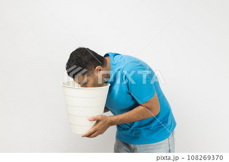 Side view portrait of sick unshaven man wearing blue T- shirt standing suffering stomachache, feels nausea and vomits, holding bin in hands. Indoor studio shot isolated on gray background. Side view portrait of sick unshaven man wearing blue T- shirt standing suffering stomachache, feels nausea and vomits, holding bin in hands. Indoor studio shot isolated on gray background. 108269370