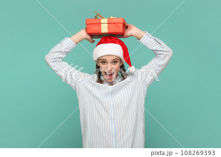 Portrait of excited amazed teenager girl with braids wearing striped shirt and Santa Claus hat, holding present box above her head, keeps mouth open. Indoor studio shot isolated on green background. Portrait of excited amazed teenager girl with braids wearing striped shirt and Santa Claus hat, holding present box above her head, keeps mouth open. Indoor studio shot isolated on green background. 108269393