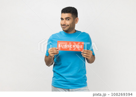 Portrait of smiling optimistic unshaven man in blue T- shirt standing holding card with subscribe inscription, looking at camera with happy expression. Indoor studio shot isolated on gray background. 108269394