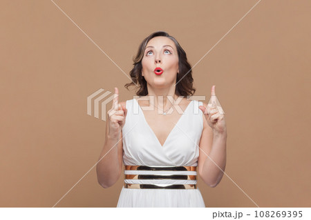 Portrait of surprised astonished middle aged woman with wavy hair standing pointing upwards at advertising area, wearing white dress. Indoor studio shot isolated on light brown background. Portrait of surprised astonished middle aged woman with wavy hair standing pointing upwards at advertising area, wearing white dress. Indoor studio shot isolated on light brown background. 108269395