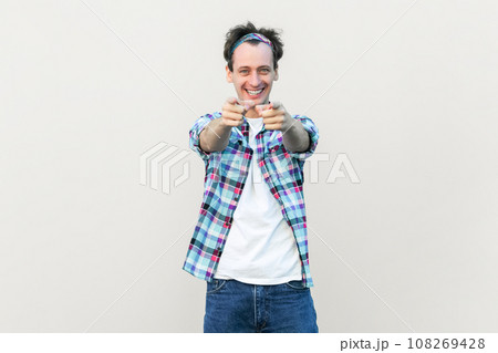 You got it. I choose you and order. Man in casual blue checkered shirt and headband points at camera with index fingers picks someone smiles broadly. Indoor studio shot isolated on gray background. 108269428