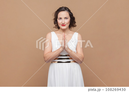 Portrait of smiling happy middle aged woman with wavy hair standing keeps palms together, looking at camera, wearing white dress. Indoor studio shot isolated on light brown background. Portrait of smiling happy middle aged woman with wavy hair standing keeps palms together, looking at camera, wearing white dress. Indoor studio shot isolated on light brown background. 108269430