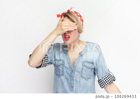 I don't want to see. Portrait of shocked scared blonde woman wearing blue denim shirt and red headband standing covering eyes with palm. Indoor studio shot isolated on gray background. 108269433