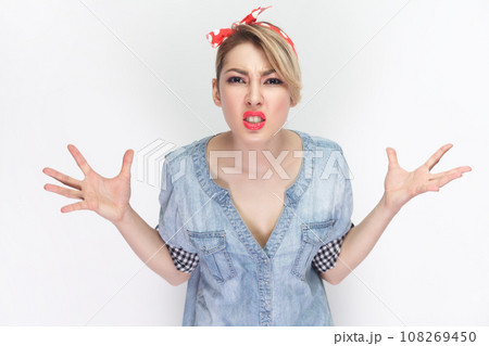 Angry annoyed woman in blue denim shirt and red headband gestures with annoyance, screams loudly, expresses irritation, reproaches partner in betrayal. Indoor studio shot isolated on gray background. Angry annoyed woman in blue denim shirt and red headband gestures with annoyance, screams loudly, expresses irritation, reproaches partner in betrayal. Indoor studio shot isolated on gray background. 108269450