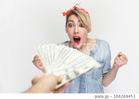 Portrait of amazed excited surprised blonde woman wearing blue denim shirt and red headband standing getting big sum of money, looks with open mouth. Indoor studio shot isolated on gray background. 108269451