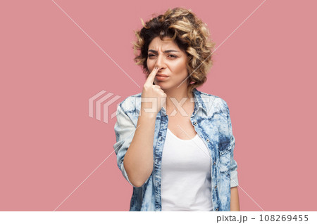You are liar. Portrait of serious beautiful young adult woman with curly hairstyle wearing blue shirt standing pointing at her nose with finger. Indoor studio shot isolated on pink background. 108269455