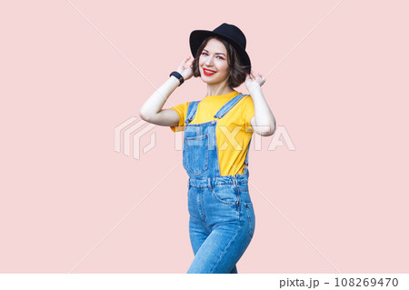 Portrait of attractive satisfied hipster woman in blue denim overalls, yellow T-shirt, keeps hands on her black hat, smiling to camera. Indoor studio shot isolated on light pink background. Portrait of attractive satisfied hipster woman in blue denim overalls, yellow T-shirt, keeps hands on her black hat, smiling to camera. Indoor studio shot isolated on light pink background. 108269470