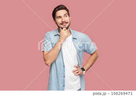 Pleasant looking bearded man standing holding chin being in good mood, imagines something nice, dreams about perfect vacation. Indoor studio shot isolated on pink background. Pleasant looking bearded man standing holding chin being in good mood, imagines something nice, dreams about perfect vacation. Indoor studio shot isolated on pink background. 108269471