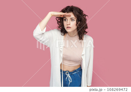 Portrait of serious attentive woman with curly hairstyle wearing casual style outfit keeps palm over forehead, looking far in future. Indoor studio shot isolated on pink background. Portrait of serious attentive woman with curly hairstyle wearing casual style outfit keeps palm over forehead, looking far in future. Indoor studio shot isolated on pink background. 108269472