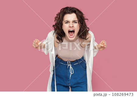 Portrait of angry woman with curly hair wearing casual style outfit standing arguing with somebody, clenched fists, screaming with hate and aggression. Indoor studio shot isolated on pink background. Portrait of angry woman with curly hair wearing casual style outfit standing arguing with somebody, clenched fists, screaming with hate and aggression. Indoor studio shot isolated on pink background. 108269473