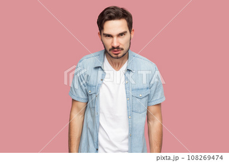 Portrait of bearded man in blue casual style shirt standing frowning, having grumpy angry look, dissatisfied with misbehavior of somebody. Indoor studio shot isolated on pink background. Portrait of bearded man in blue casual style shirt standing frowning, having grumpy angry look, dissatisfied with misbehavior of somebody. Indoor studio shot isolated on pink background. 108269474