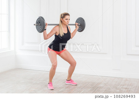 Side view portrait of sporty athletic beautiful woman in pink shorts and black top doing squats and exercising at the gym with the barbell on white wall background. Indoor studio shot. Side view portrait of sporty athletic beautiful woman in pink shorts and black top doing squats and exercising at the gym with the barbell on white wall background. Indoor studio shot. 108269493
