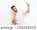 Portrait of joyful handsome playful bearded man wearing striped t-shirt standing making selfie, showing v sign to his followers. Indoor studio shot isolated on gray background. 108269502