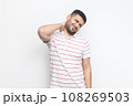 Portrait of handsome sick tired bearded man wearing striped t-shirt standing massaging his painful neck, frowning face from terrible pain. Indoor studio shot isolated on gray background. 108269503