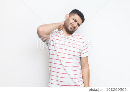 Portrait of handsome sick tired bearded man wearing striped t-shirt standing massaging his painful neck, frowning face from terrible pain. Indoor studio shot isolated on gray background. Portrait of handsome sick tired bearded man wearing striped t-shirt standing massaging his painful neck, frowning face from terrible pain. Indoor studio shot isolated on gray background. 108269503