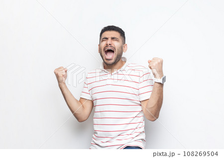 Portrait of extremely happy joyful cheerful bearded man wearing striped t-shirt standing with clenched raised fists, screaming, celebrating victory. Indoor studio shot isolated on gray background. Portrait of extremely happy joyful cheerful bearded man wearing striped t-shirt standing with clenched raised fists, screaming, celebrating victory. Indoor studio shot isolated on gray background. 108269504