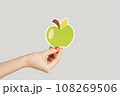 Closeup of woman hand showing paper green apple, symbol of healthy eating. Indoor studio shot isolated on gray background. 108269506