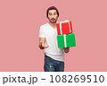 Portrait of puzzled confused bearded man in white T-shirt and beany hat holding two present boxes in hands, looking at camera with surprised expression. Indoor studio shot isolated on pink background 108269510