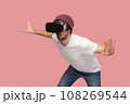 Portrait of amazed wary bearded man in white T-shirt and beany hat, wearing vr, playing in headset, trying to avoid monster in video game. Indoor studio shot isolated on pink background. 108269544