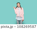 Portrait of excited smiling positive teenager girl with braids wearing pink jacket pointing index finger away, showing copy space for advertisement. Indoor studio shot isolated on green background. 108269587