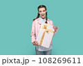 Portrait of excited positive teenager girl wearing pink jacket standing wrapped present box in hands, looking at camera, waiting holiday to give gift. Indoor studio shot isolated on green background. 108269611