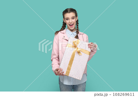 Portrait of excited positive teenager girl wearing pink jacket standing wrapped present box in hands, looking at camera, waiting holiday to give gift. Indoor studio shot isolated on green background. Portrait of excited positive teenager girl wearing pink jacket standing wrapped present box in hands, looking at camera, waiting holiday to give gift. Indoor studio shot isolated on green background. 108269611