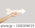 Closeup of woman hand holding white arrow pointing away, showing direction aside. Indoor studio shot isolated on gray background. 108269619