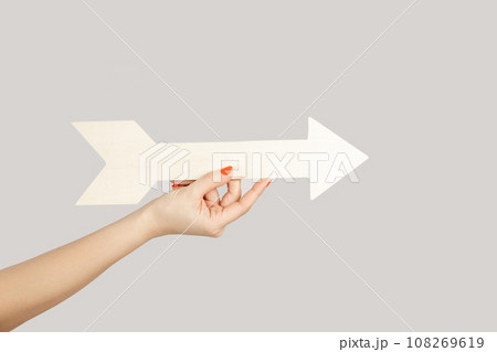Closeup of woman hand holding white arrow pointing away, showing direction aside. Indoor studio shot isolated on gray background. 108269619