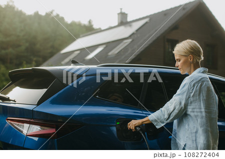 Portrait of a woman charging electric car in front of her house with solar panels on roof. Mother plugging the charger into the charging port. Solar energy and sustainable lifestyle of young family. 108272404