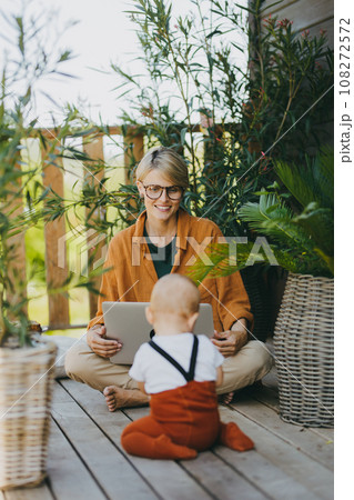 Mother holding baby while working on laptop in garden. Businesswoman working remotely from outdoor home office and taking care of little son. Life work balance with kid. Mother holding baby while working on laptop in garden. Businesswoman working remotely from outdoor home office and taking care of little son. Life work balance with kid. 108272572