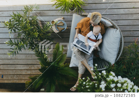 Top view of mother holding baby while working on laptop in garden. Businesswoman working remotely from outdoor home office and taking care of little son. Life work balance with kid. Top view of mother holding baby while working on laptop in garden. Businesswoman working remotely from outdoor home office and taking care of little son. Life work balance with kid. 108272585